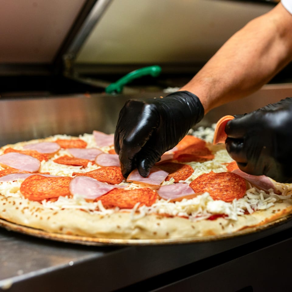 Gloved hands arranging pepperoni and ham on a Numero Uno Pizza before baking.