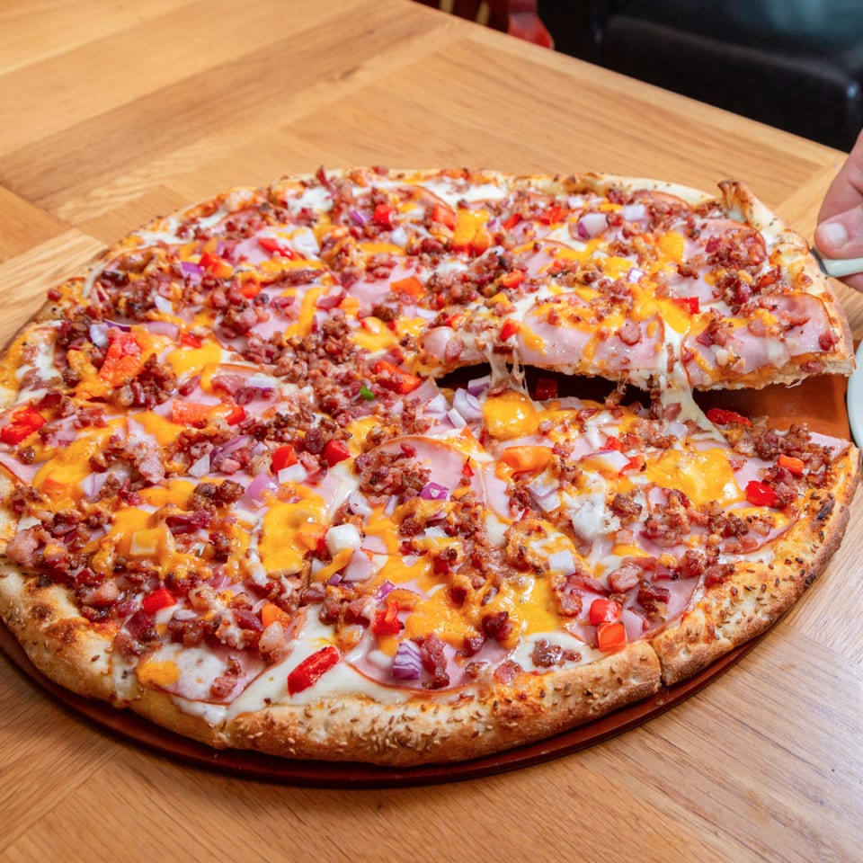 A hand lifting a slice of Numero Uno Pizza with melted cheese, ham, bacon, red peppers, and onions.