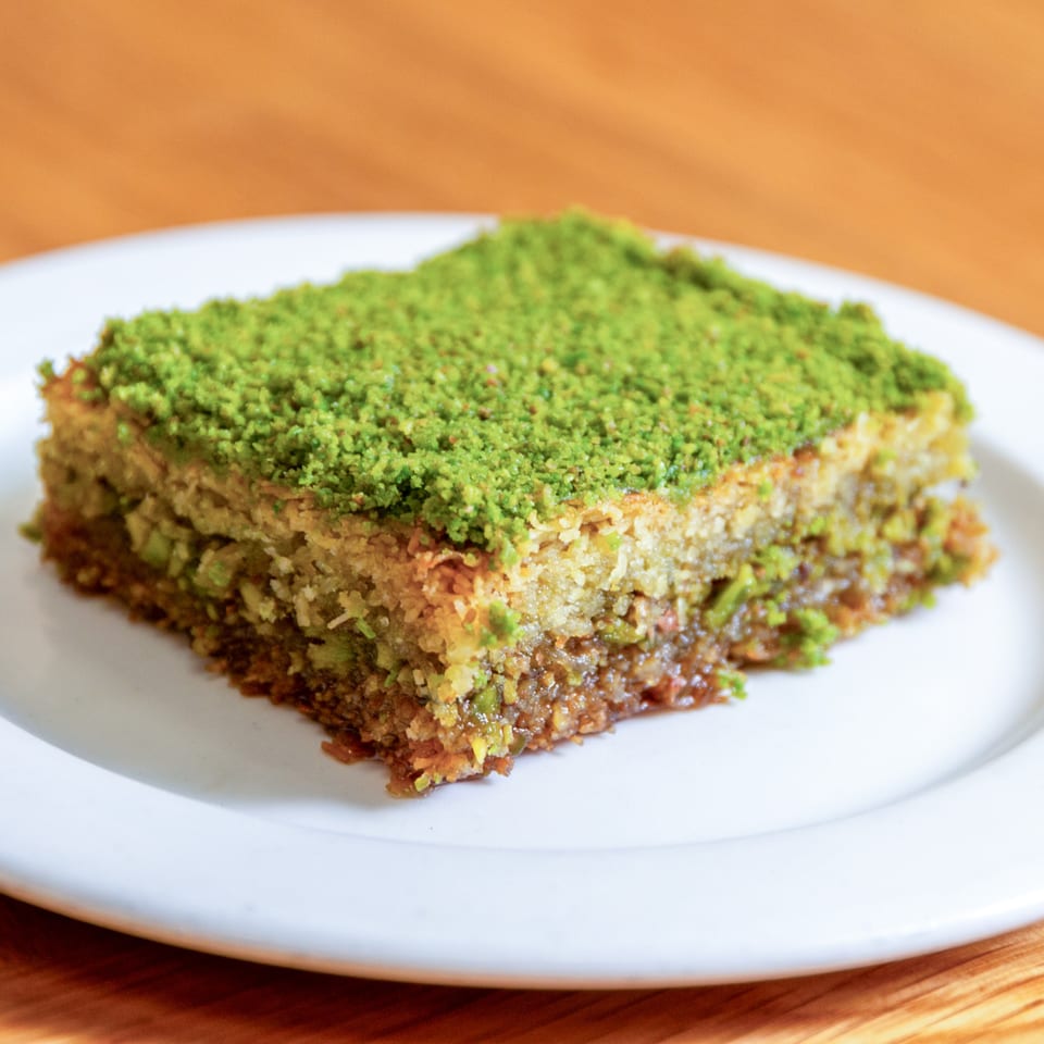 A square piece of pistachio baklava on a white plate, with a wooden table background.