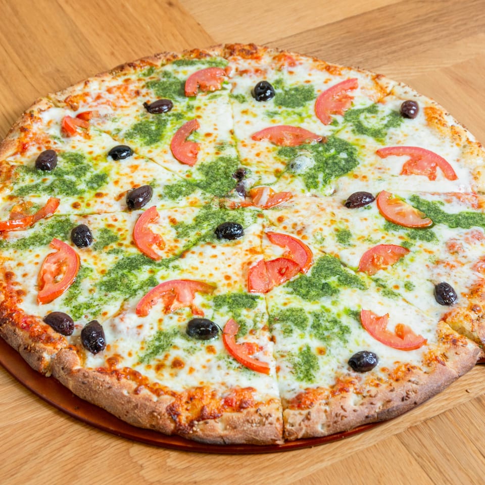 Numero Uno Pizza with pesto, tomatoes, and black olives on a wooden table.