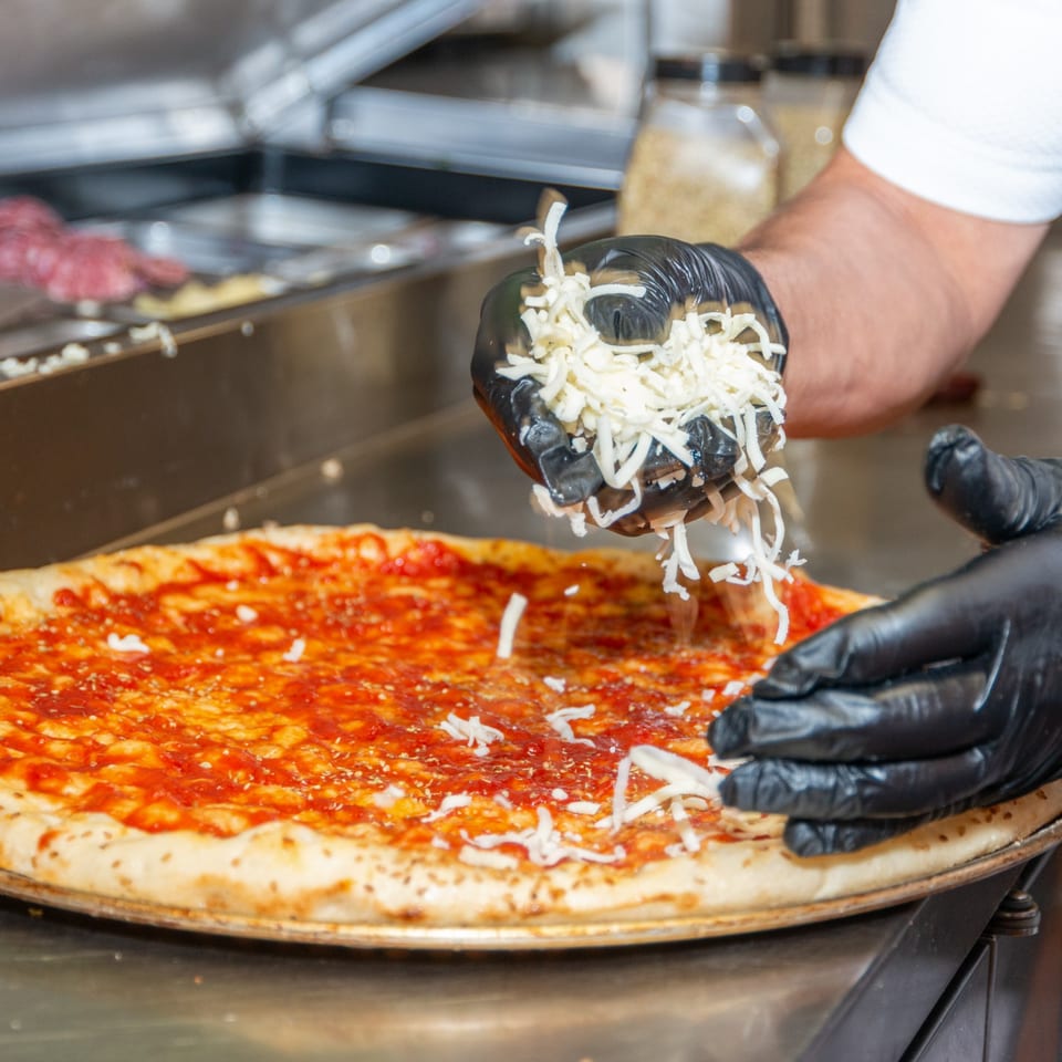 Chef's gloved hands sprinkle shredded mozzarella cheese onto a Numero Uno Pizza with red sauce.